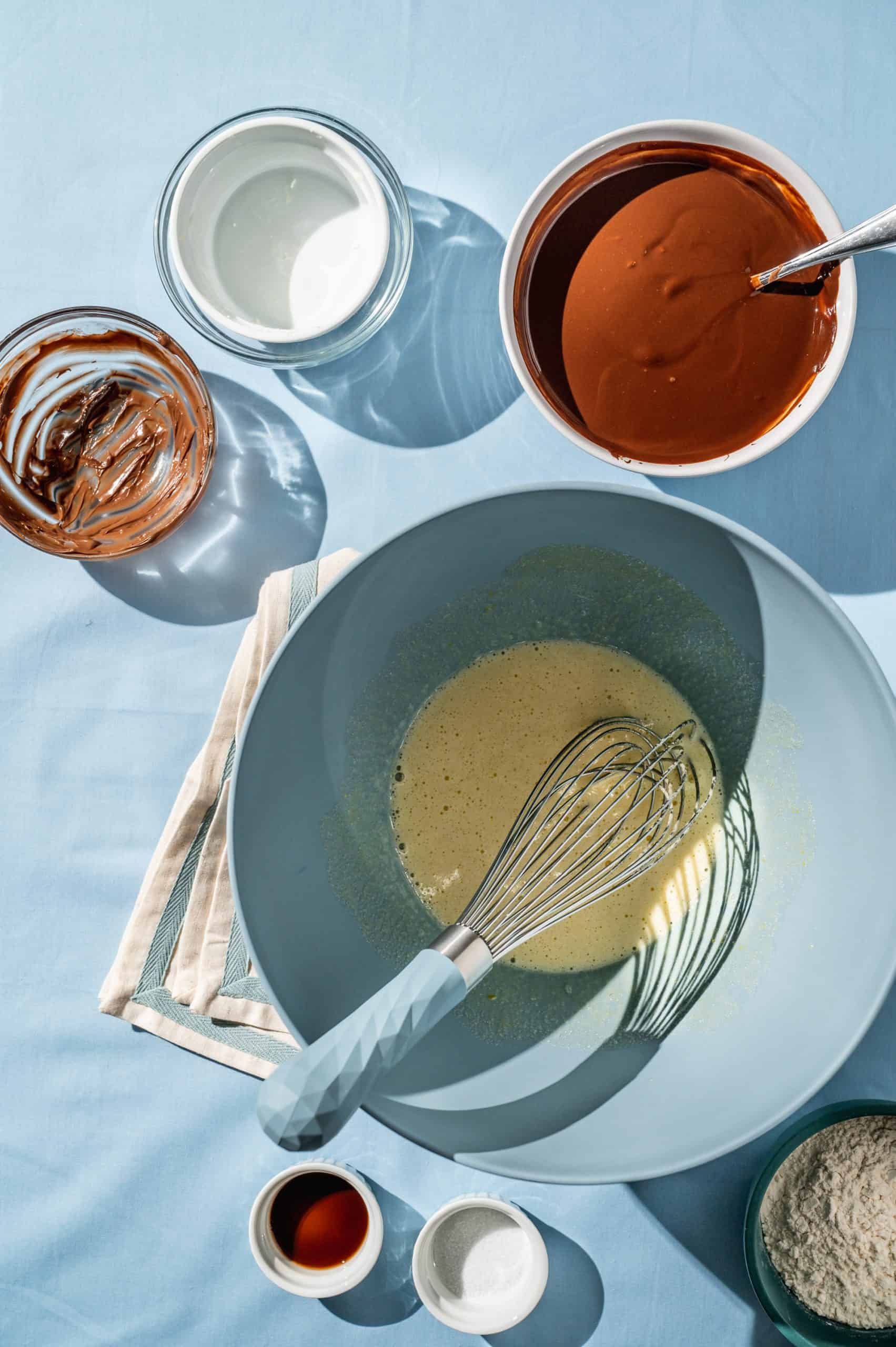 large mixing bowl with eggs and sugar with a blue handled whisk, bowl of melted chocolate to the side