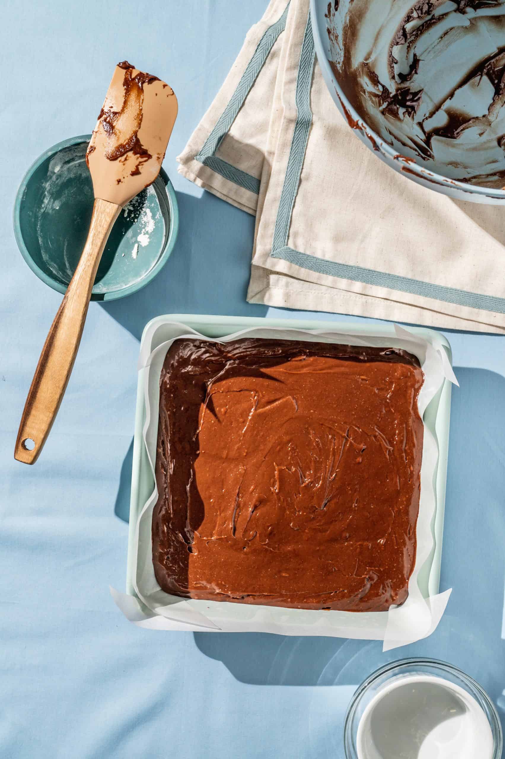 brownie batter in a square baking pan on a blue tablecloth