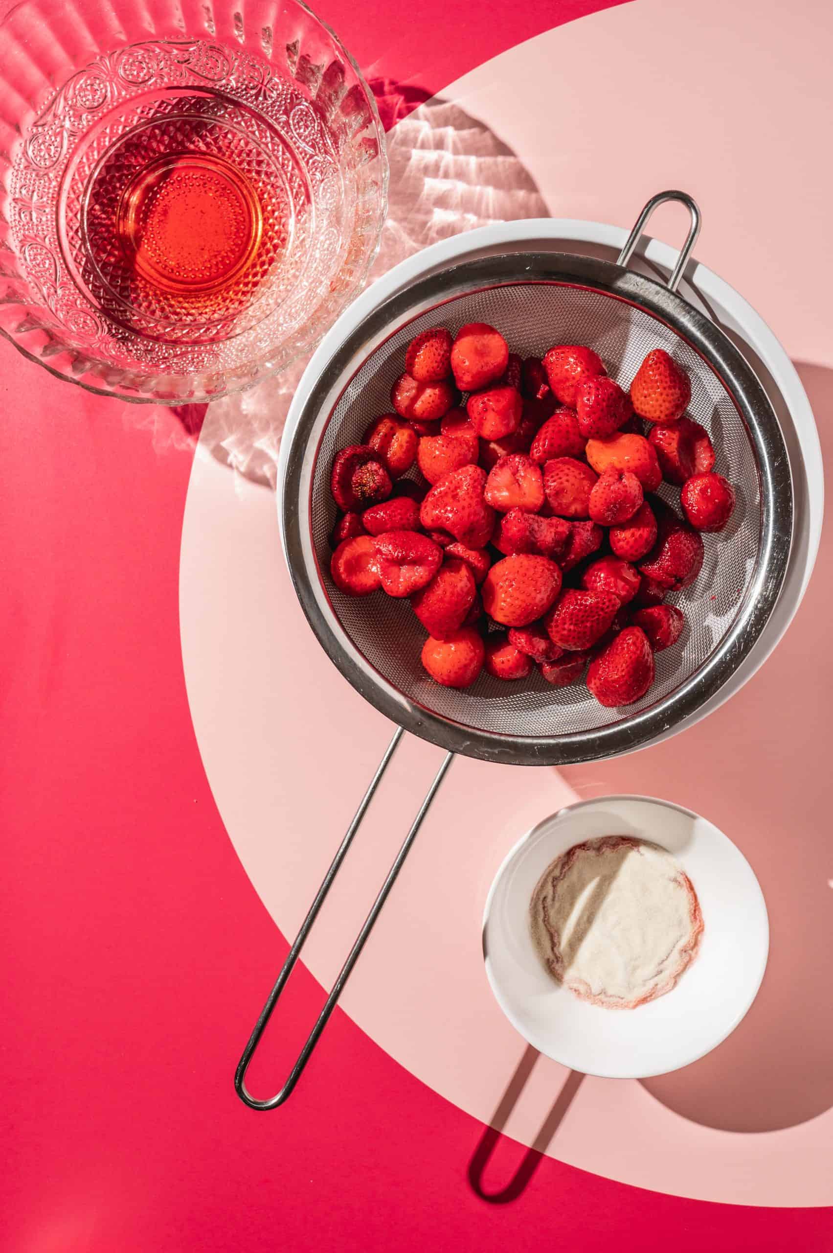 frozen strawberries in a strainer set over a bowl with a small bowl of gelatin on the side