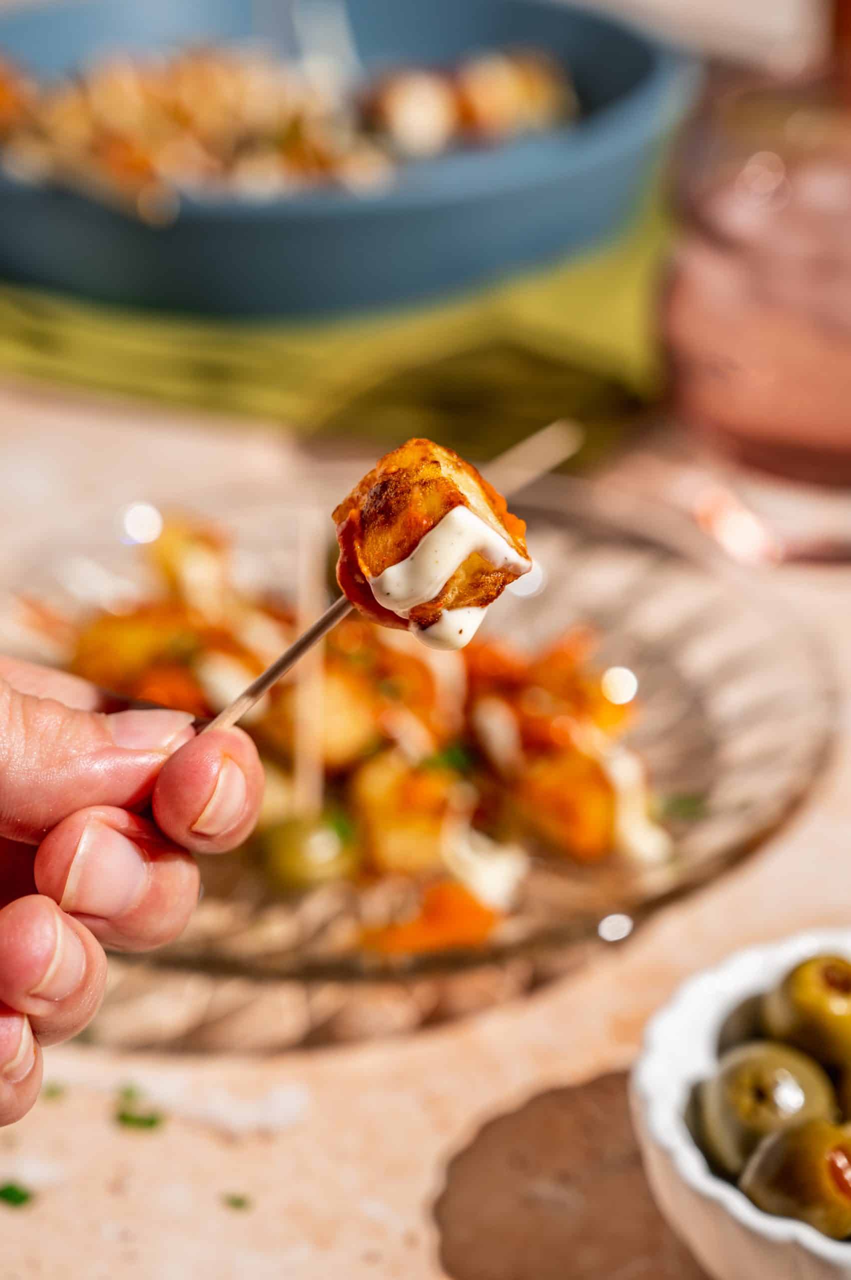 hand holding a toothpick with a crispy potato on it, drizzled with aioli and bravas sauce