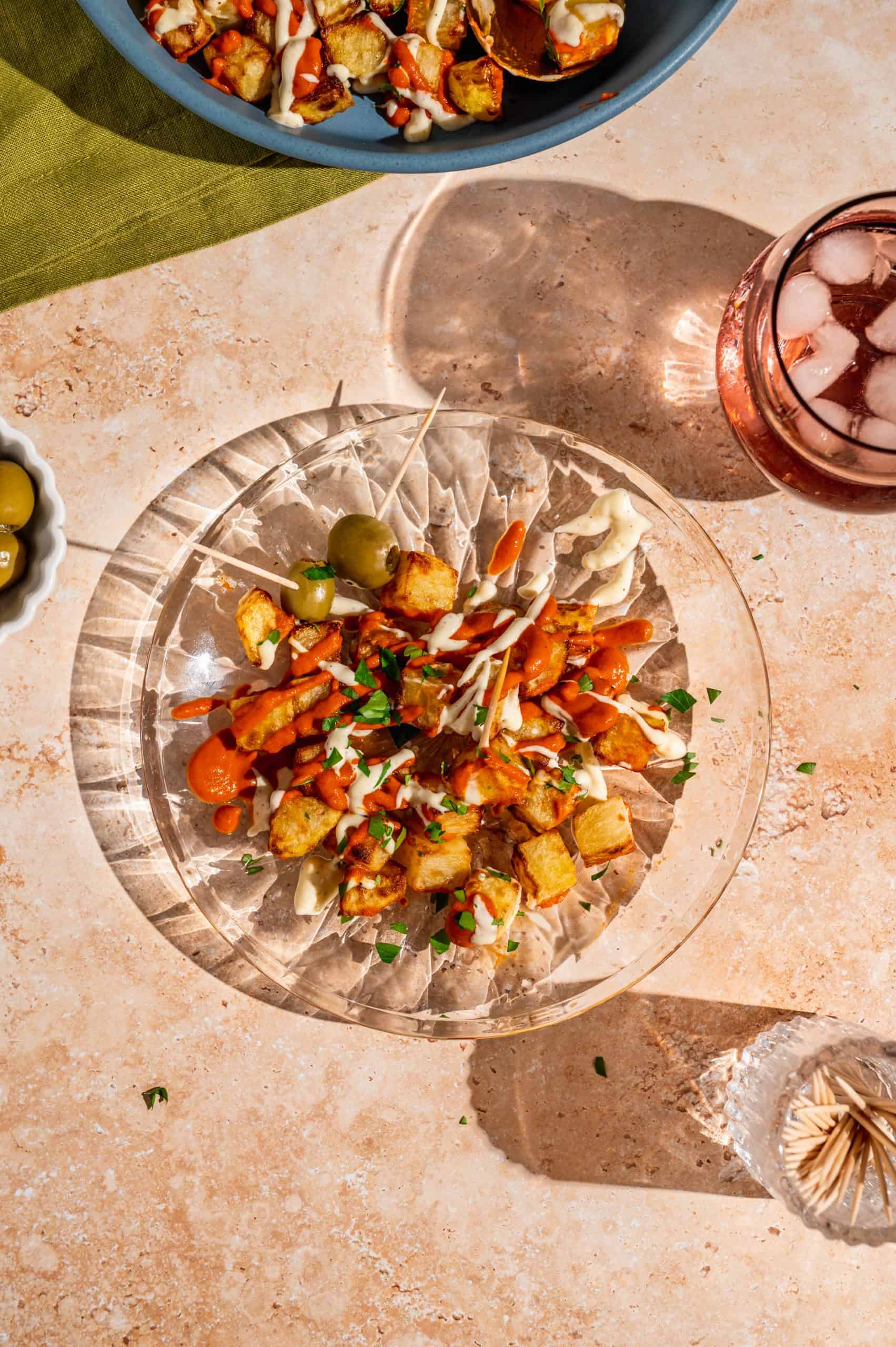 a portion of patatas bravas and some green olives on toothpicks on a small glass plate