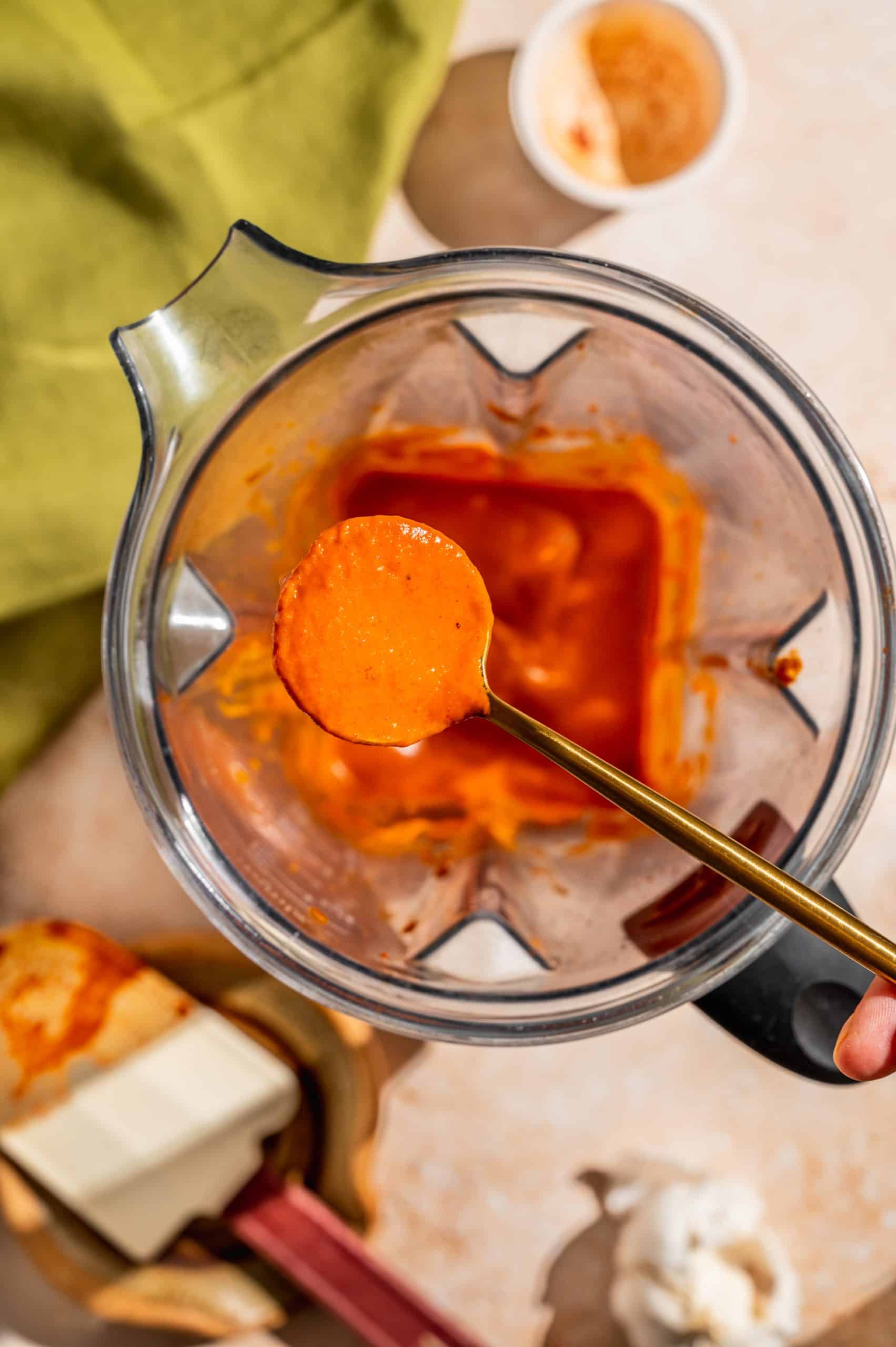 gold spoon holding smooth red-orange sauce over a blender