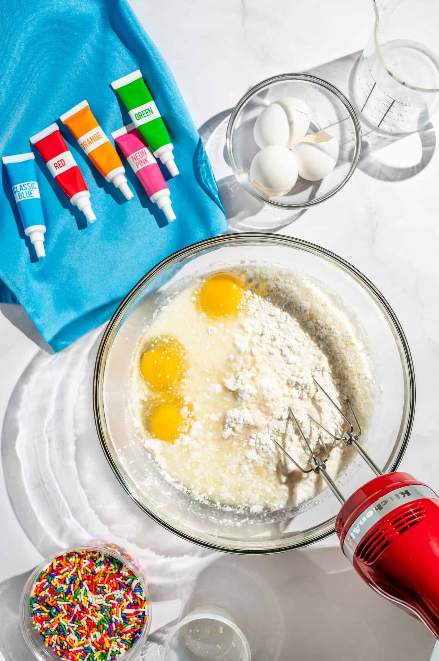 glass mixing bowl with vanilla cake mix, 3 eggs, oil, and milk being mixed with a handheld electric mixer