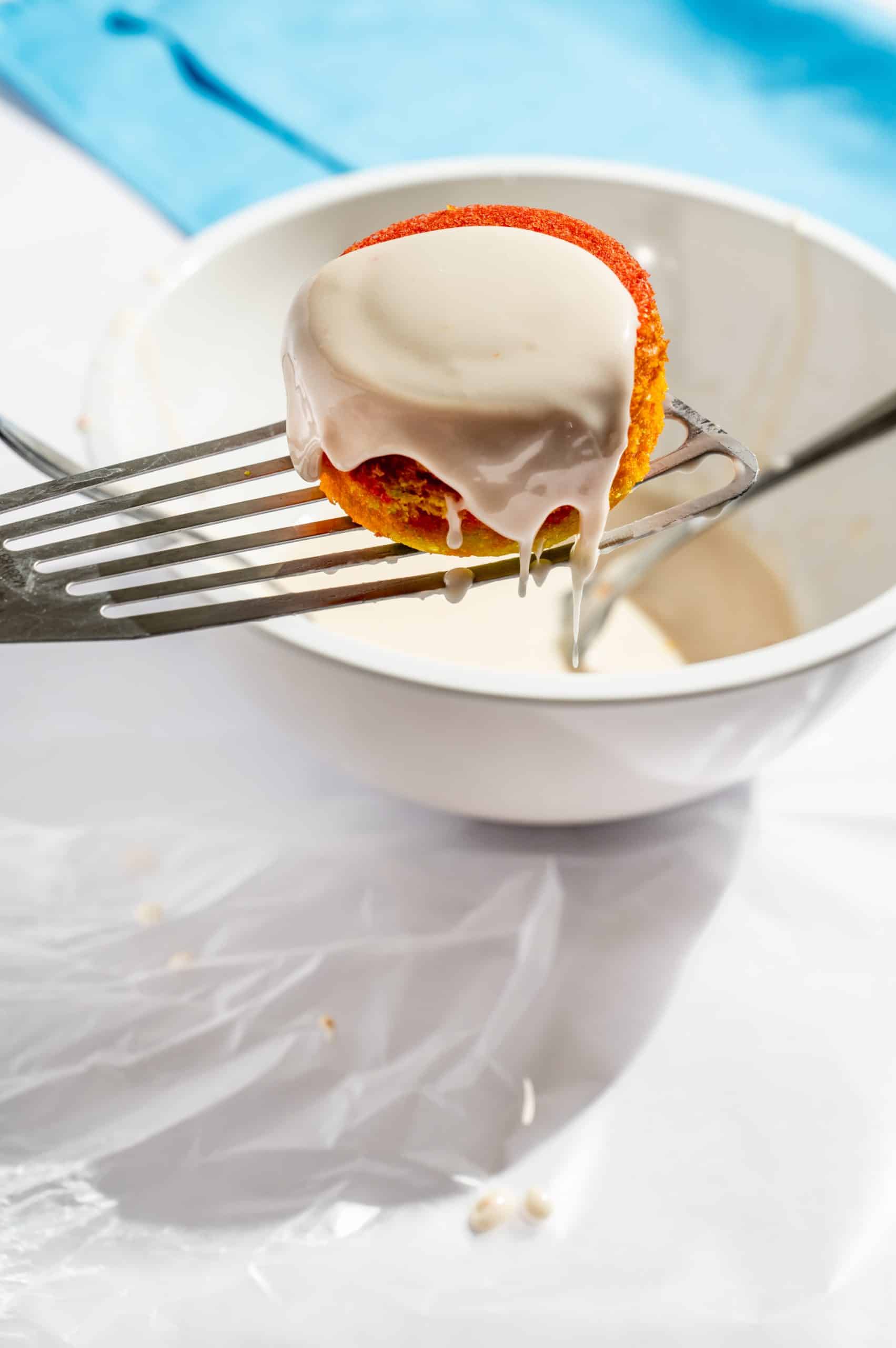 dipping rainbow cupcake in vanilla glaze allowing excess to drip off back into the bowl