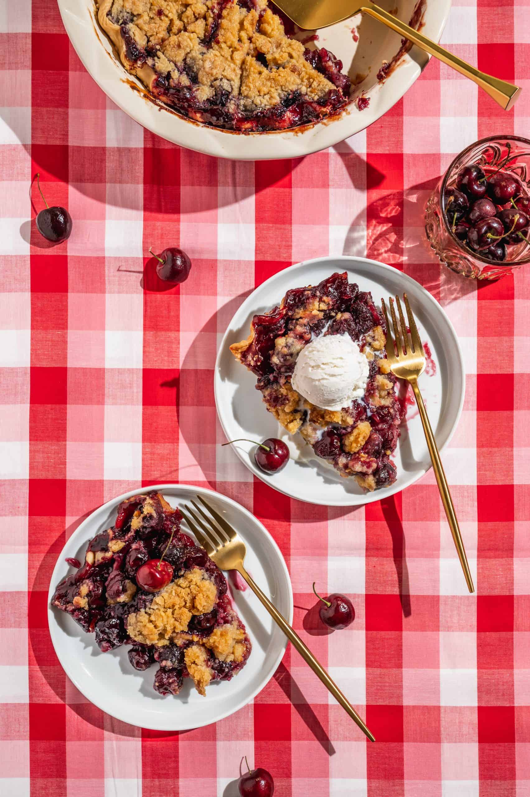 two slices on cherry crumble pie on white plates with gold forks on a checkered red tablecloth