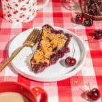 slice of cherry crumble pie on a white plate with fresh cherries on the side