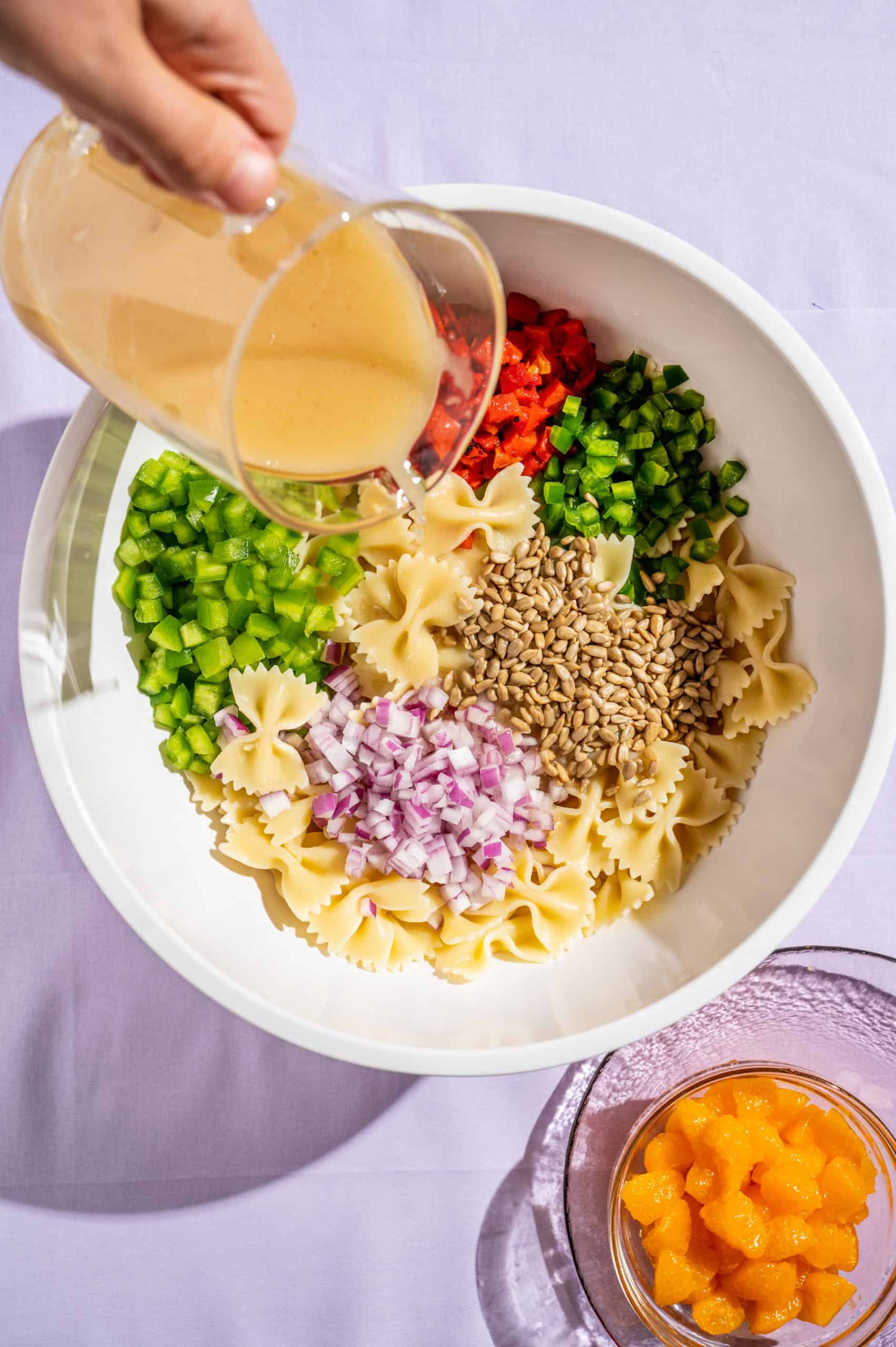 Pouring honey vinaigrette over bowl with pasta and prepped vegetables, with small bowl of oranges on the side.