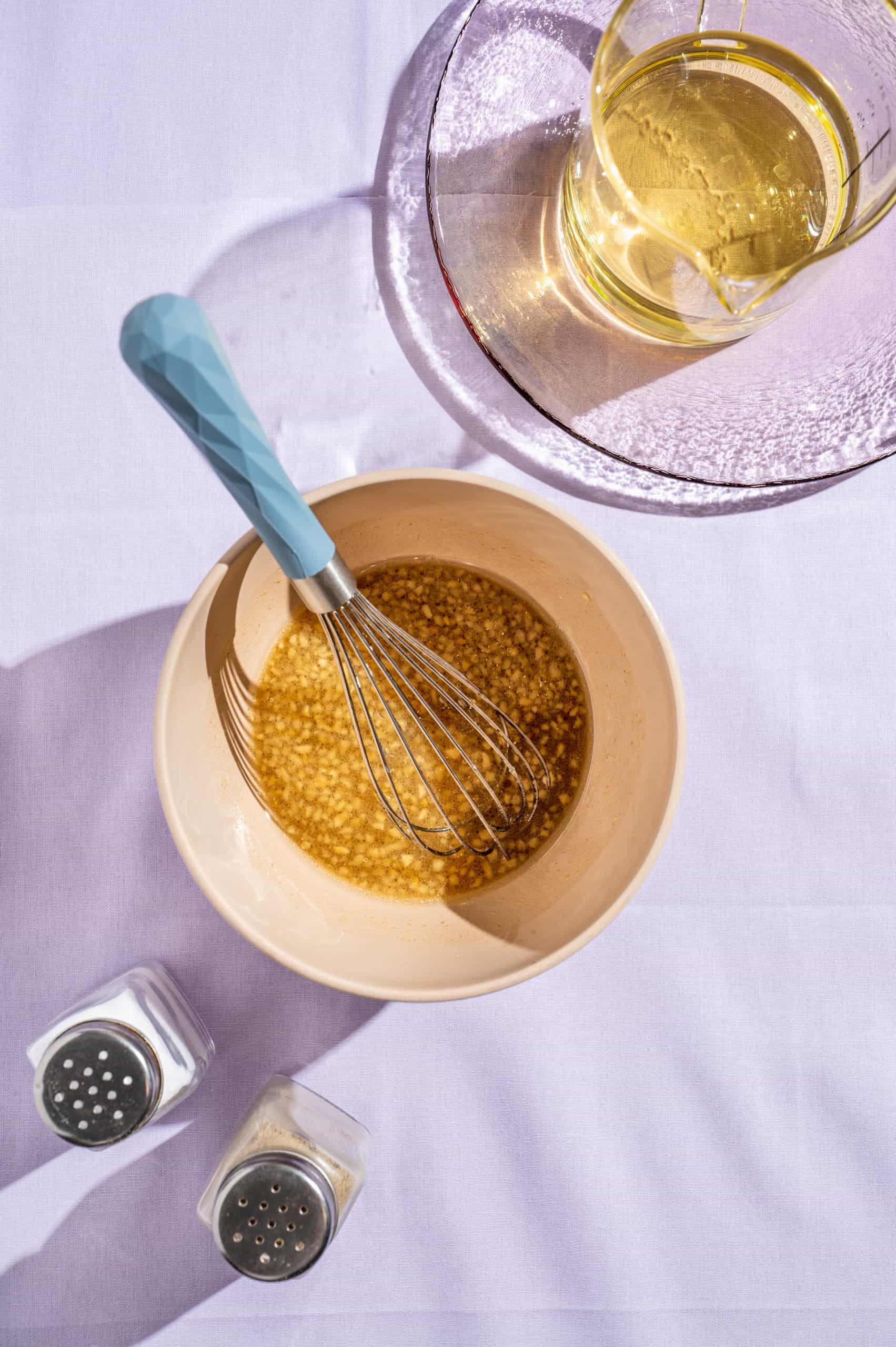 Honey garlic vinaigrette in a small mixing bowl with a whisk, extra oil in a liquid measuring cup on the side.