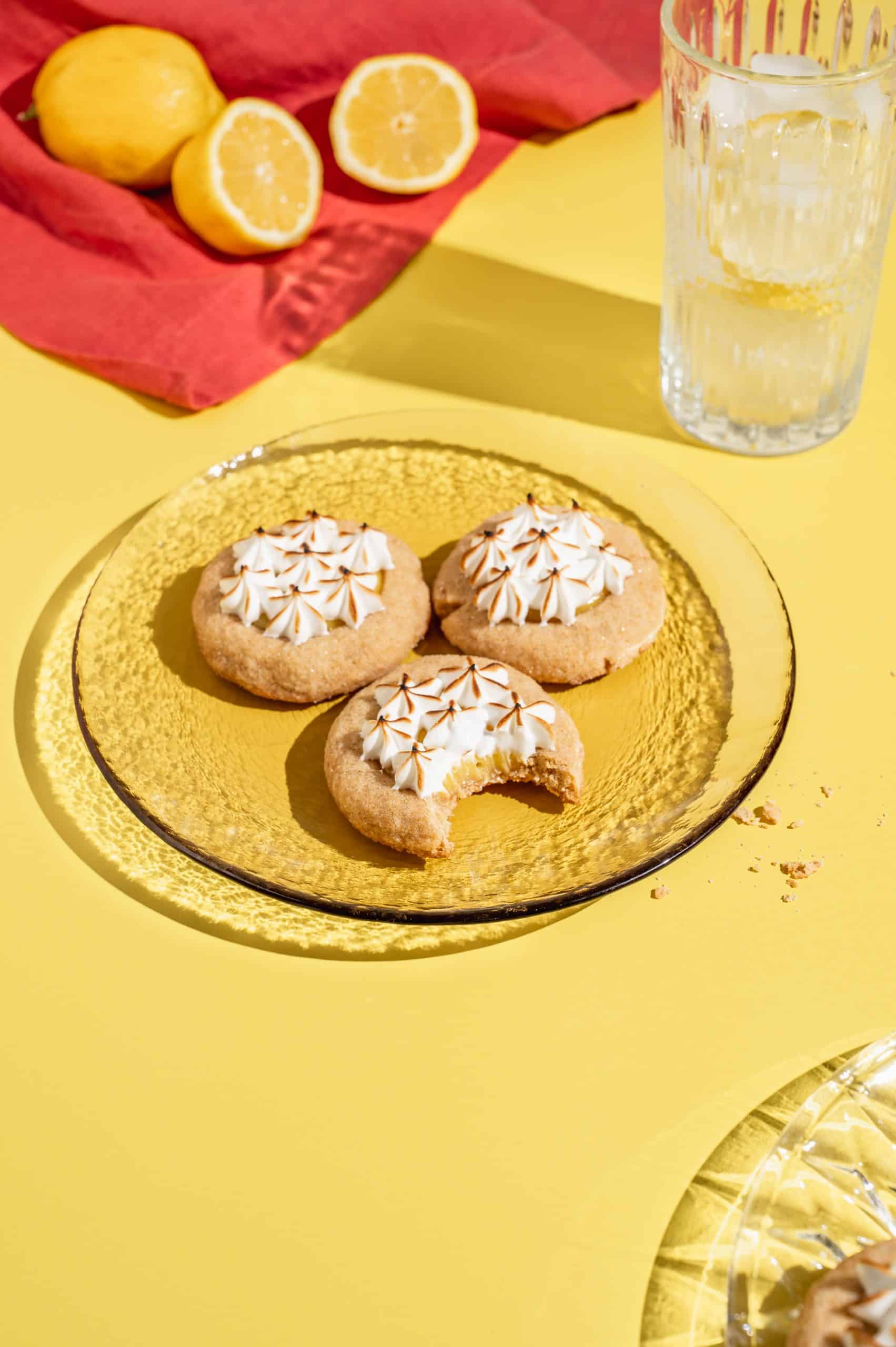 3 lemon meringue pie cookies on a glass plate with lemons in the background