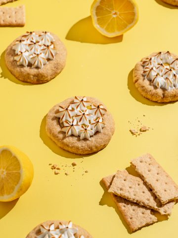 lemon meringue cookies on a yellow surface surrounded by lemon halves and graham cracker pieces
