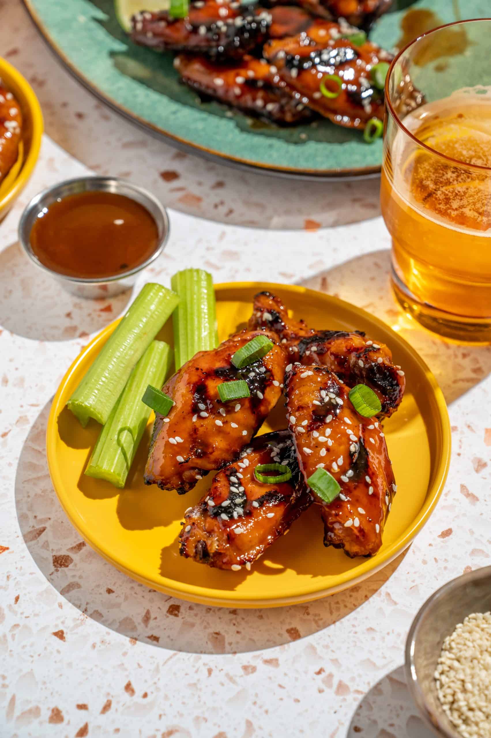 Small plate with Asian grilled chicken wings and celery, a glass of beer and extra sauce on the side, with a platter in the background