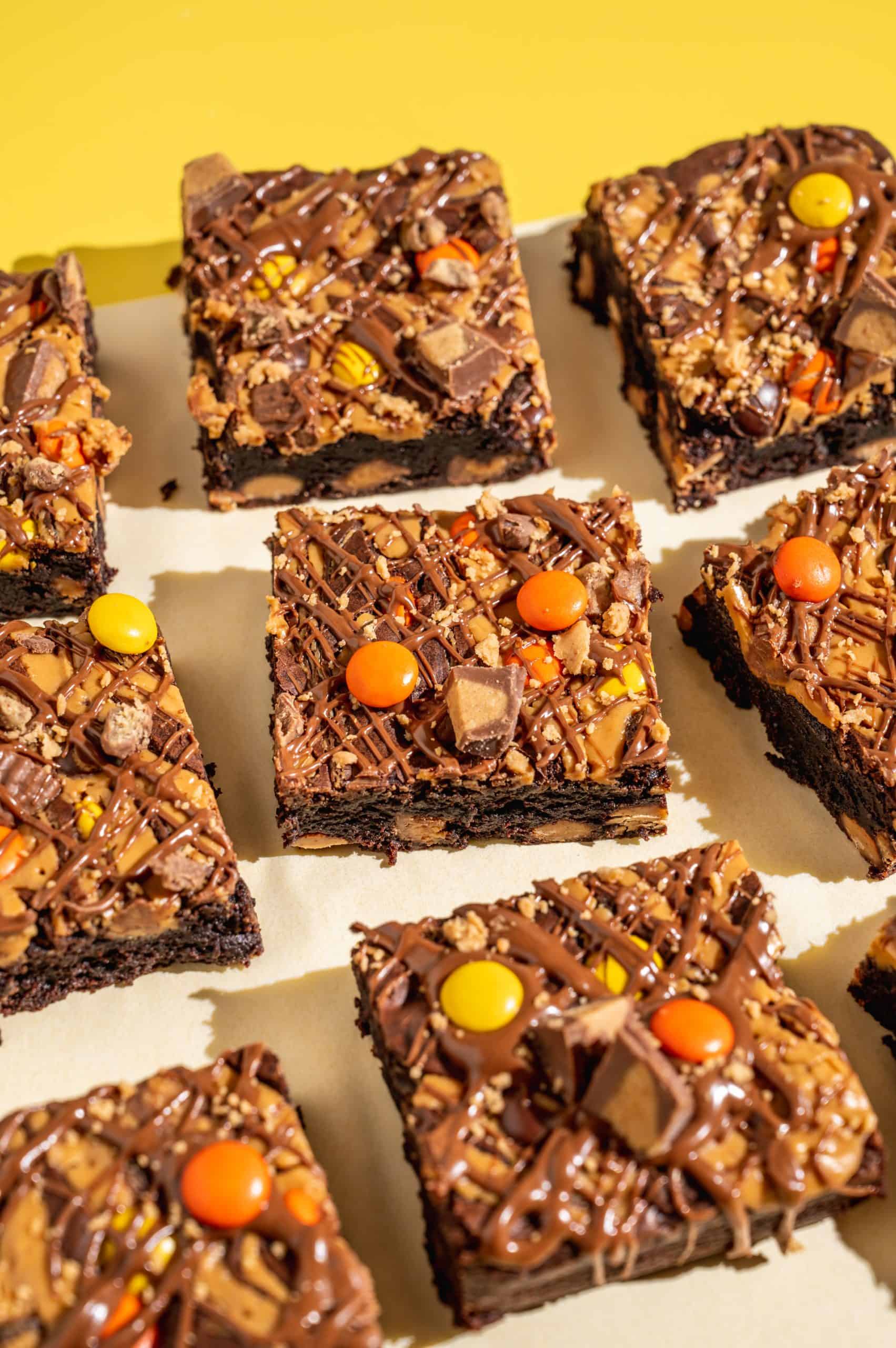 Nine square Reeses Pieces brownies on white parchment over a bright yellow surface.