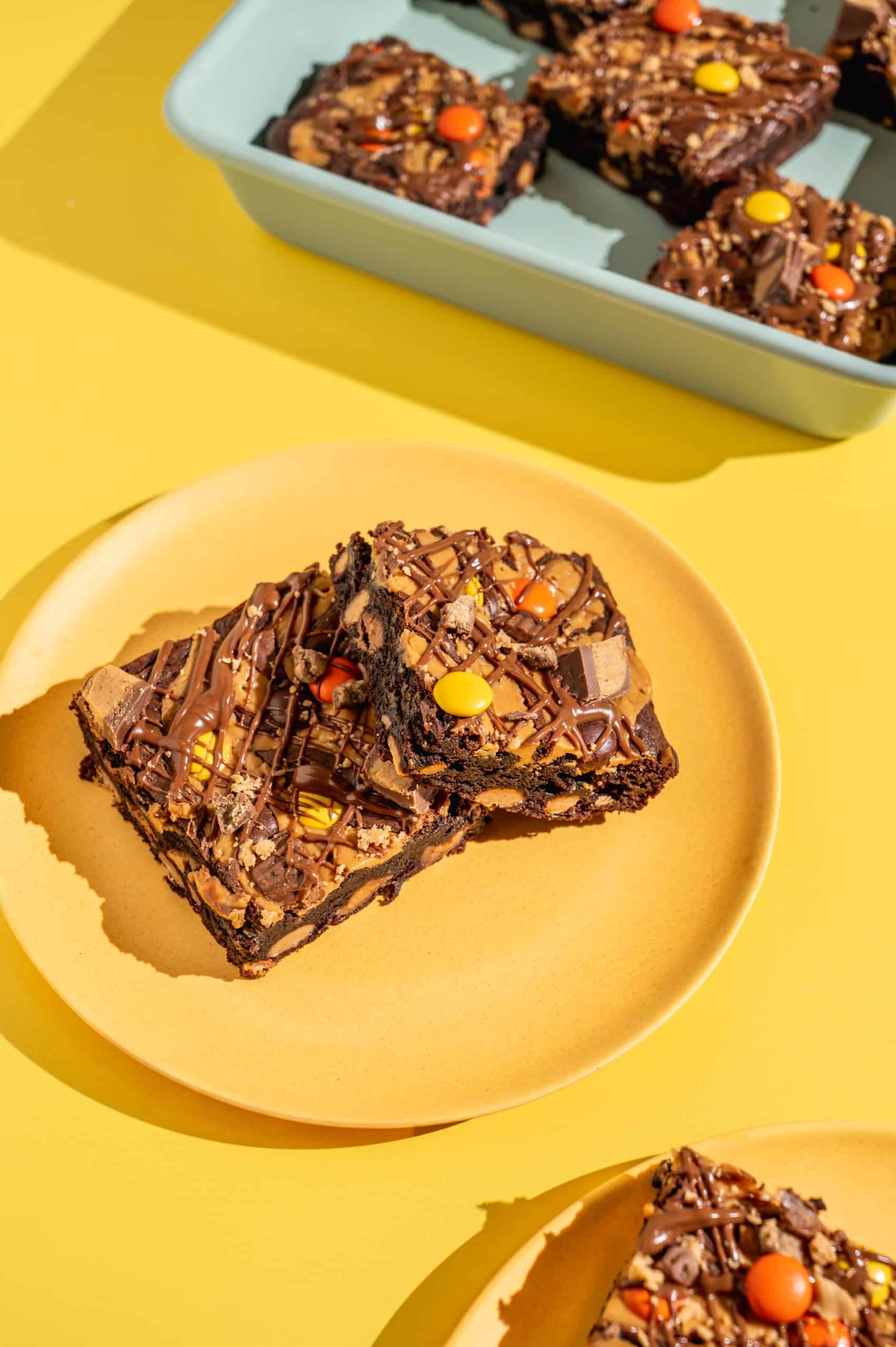 Two Reese's Pieces brownies on a yellow plate with extra brownies in the pan in the background.