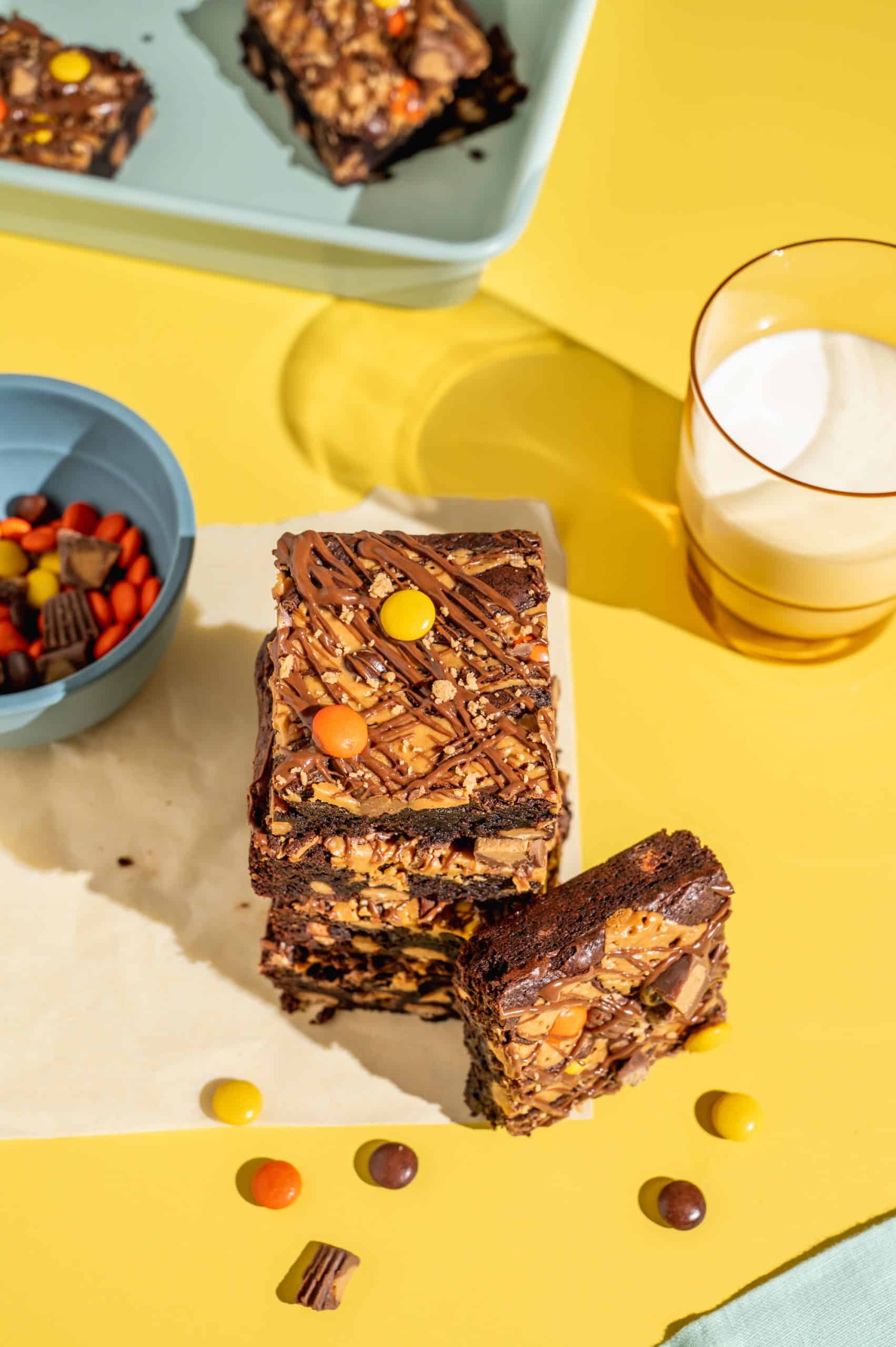 A stack of 4 Reese's brownies with a few brownies and a glass of milk on the side.