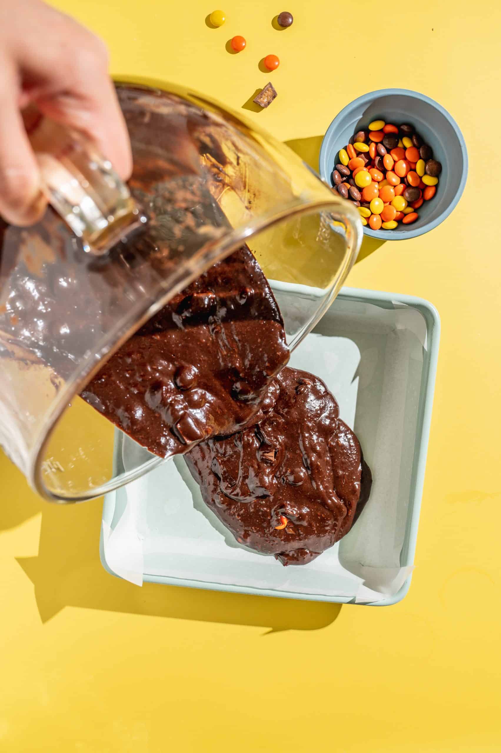 Pouring Reeses pieces brownie batter into a square baking pan lined with parchment paper.