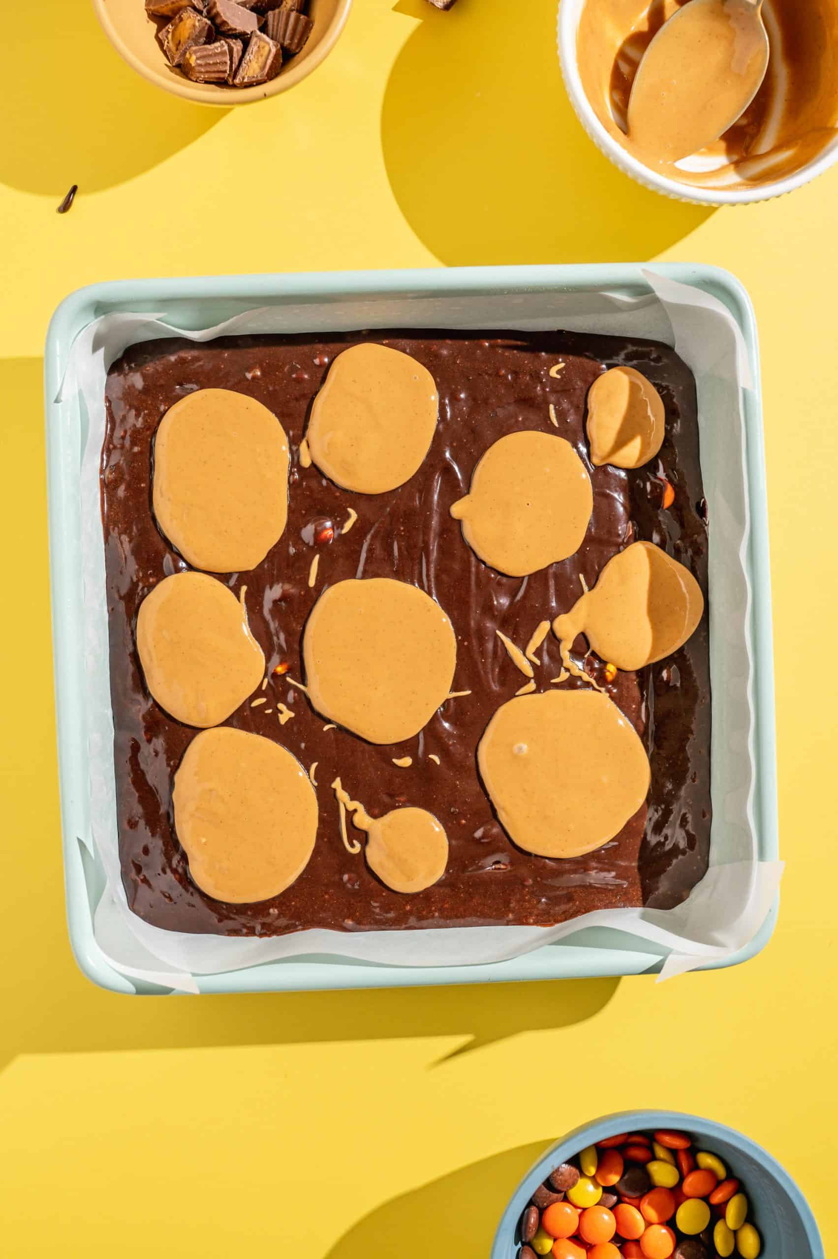 Brownie batter in a square baking pan topped with ten dollops of melty peanut butter.